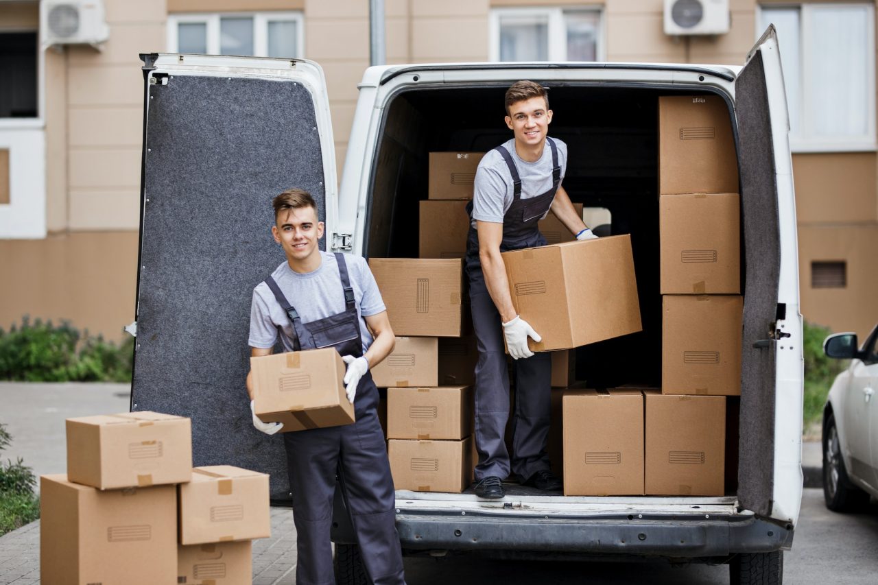 due giovani traslocatori sorridenti che indossano uniformi stanno scaricando il furgone pieno di scatole per il trasloco casa.jpg